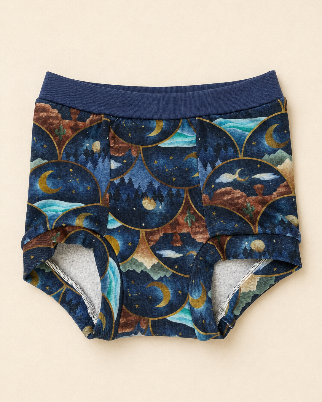 A pair of children's briefs with a navy blue elastic waistband and a detailed celestial pattern featuring crescent moons, stars, and mountains inside scalloped arches. The inner lining is light gray.