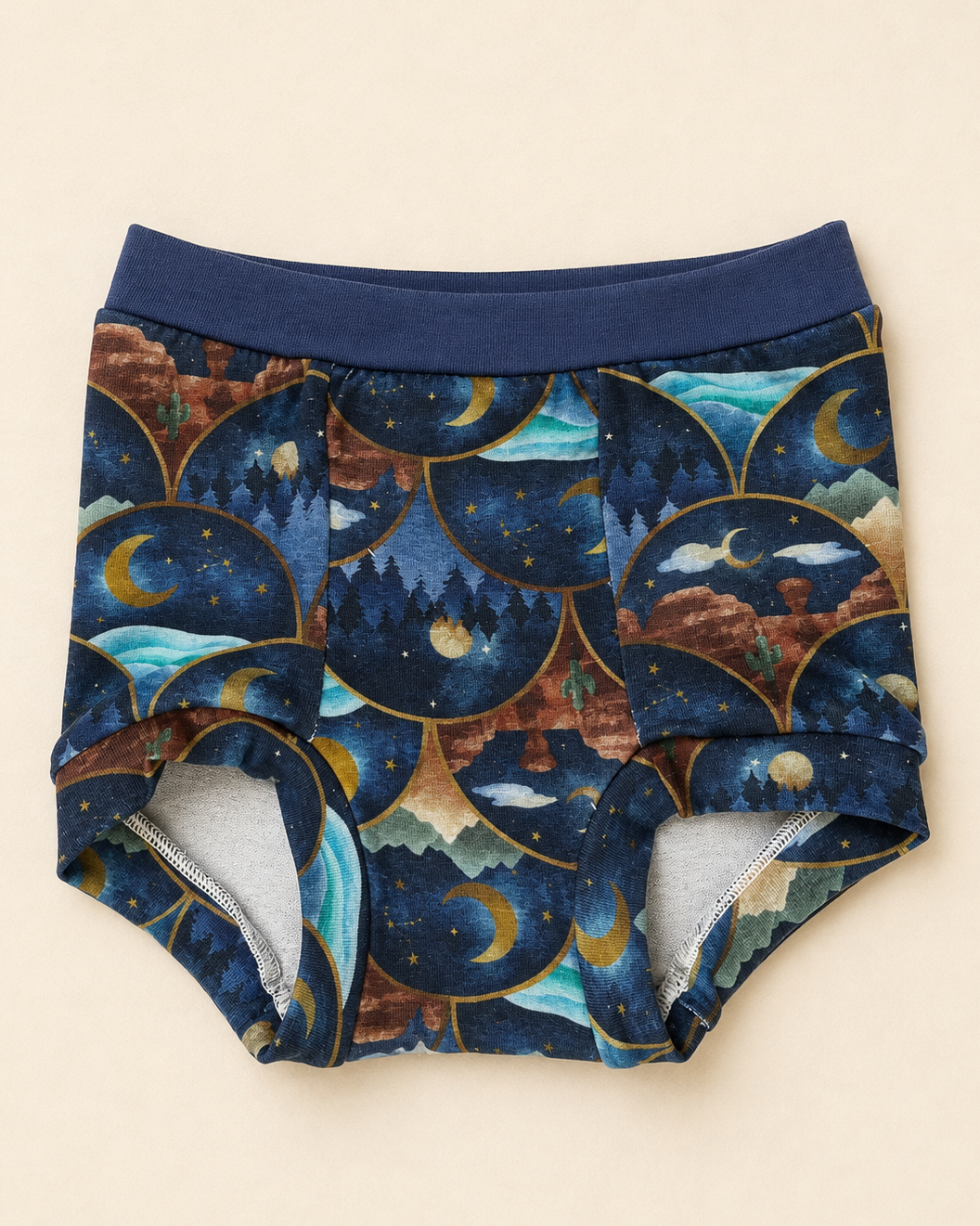 A pair of children's briefs with a navy blue elastic waistband and a detailed celestial pattern featuring crescent moons, stars, and mountains inside scalloped arches. The inner lining is light gray.