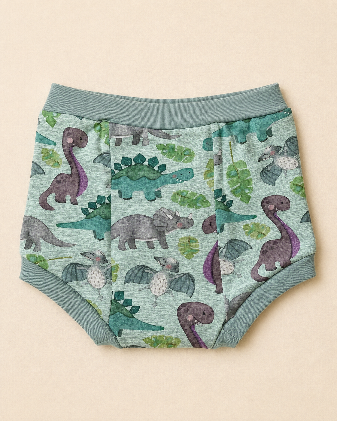 A pair of children's diaper cover or underwear with a dinosaur print, featuring a sage green waistband and leg cuffs. The fabric has an all-over pattern of various dinosaurs in purple, green, and gray on a light mint green background, laid flat on a light beige background.