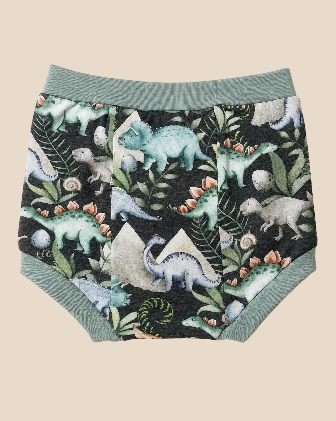 A pair of children's diaper cover featuring a dinosaur and prehistoric plant pattern on a black background, with sage green trim and waistband, laid flat on a beige background.