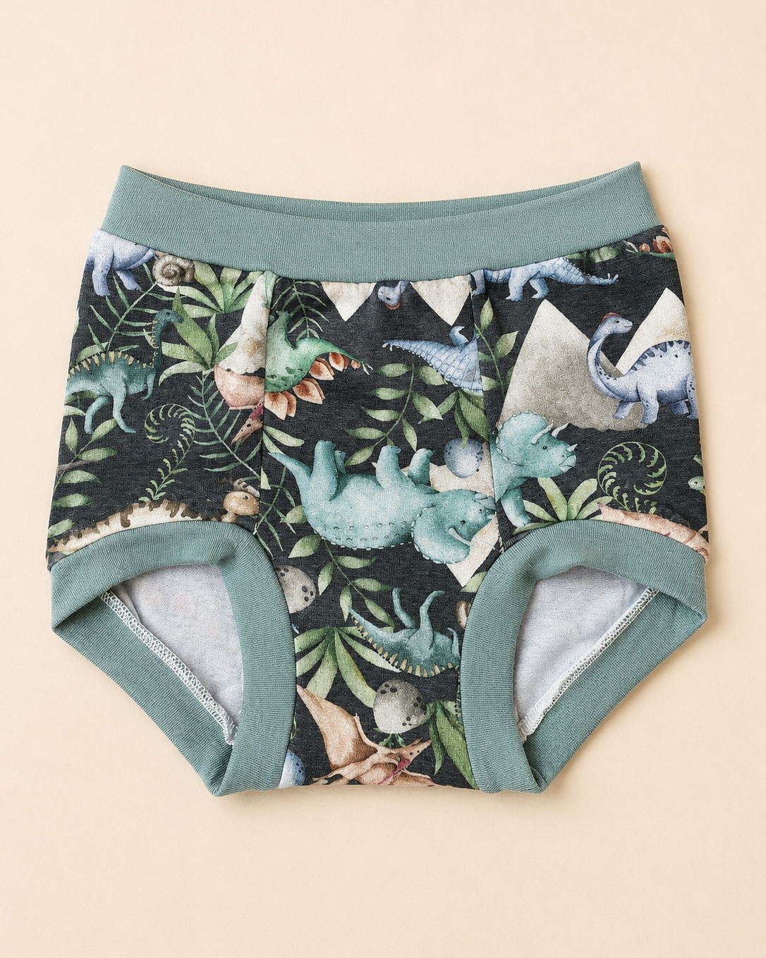 A pair of toddler boy's underwear with a dark green and black dinosaur print, featuring sage green trim and lining, laid flat on a light beige background.