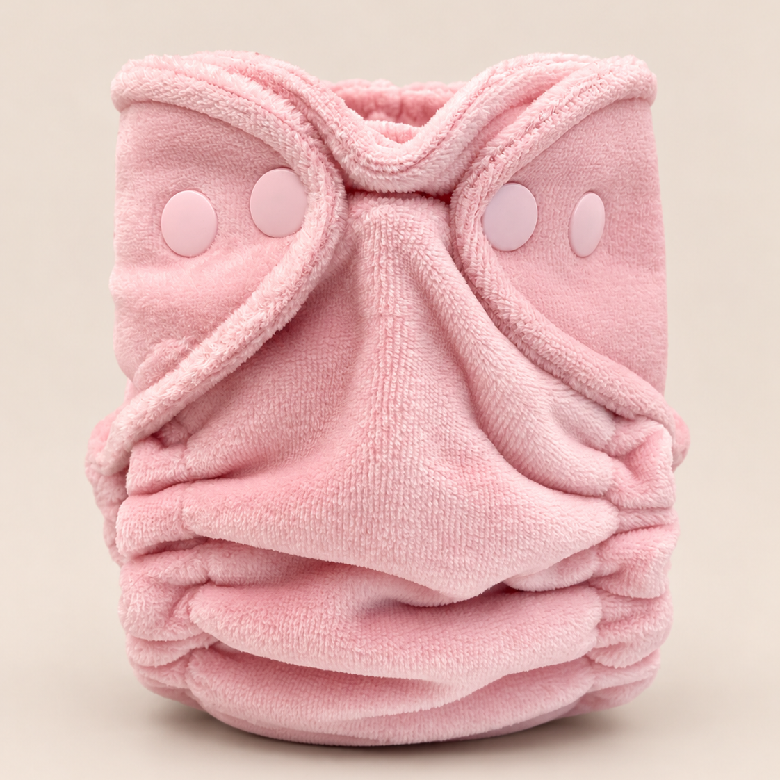 A soft pink reusable cloth diaper with a plush terry cloth texture, featuring adjustable hook-and-loop closure tabs on the front, photographed against a solid light beige background.