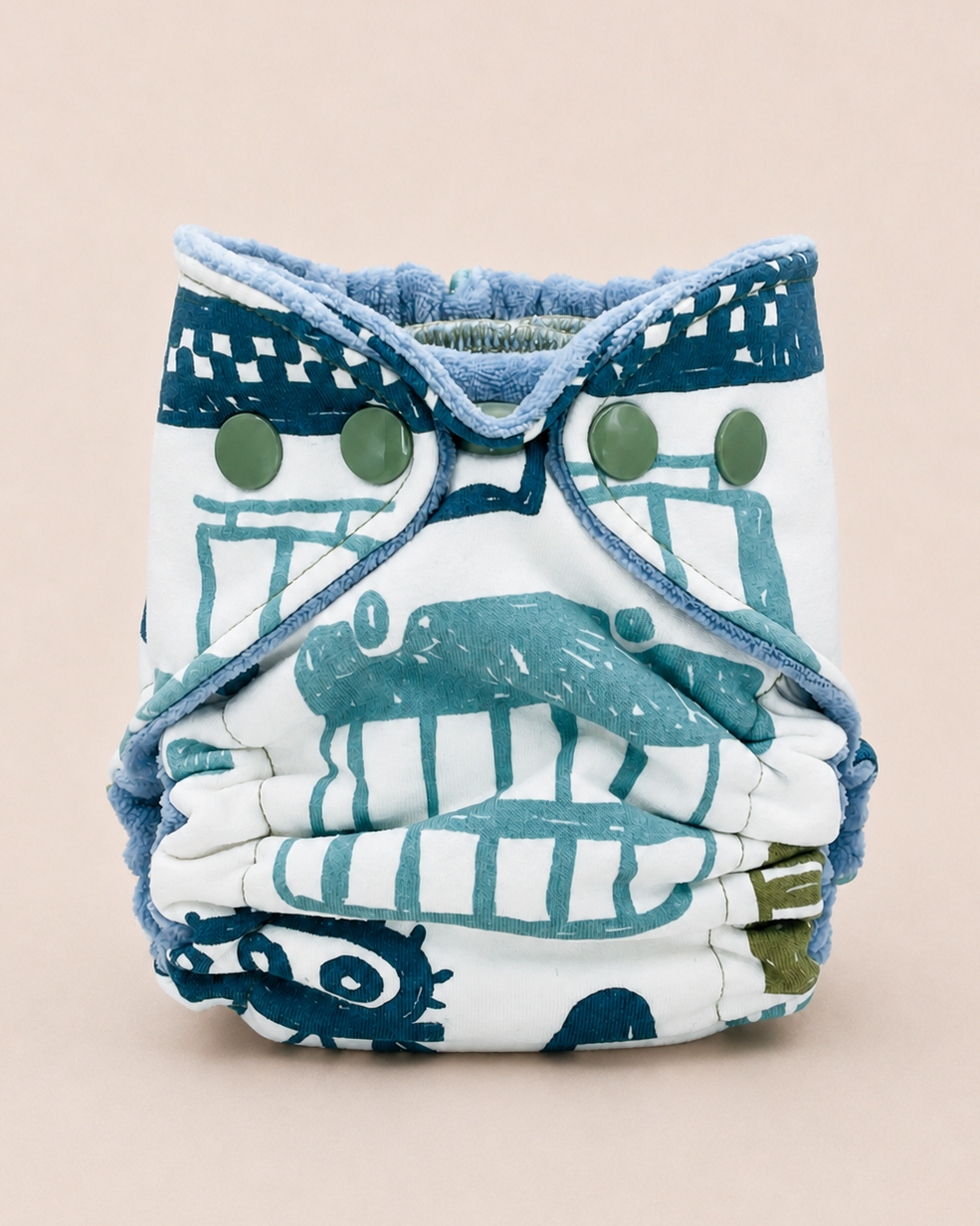 A reusable cloth diaper with a white base and a blue-green nautical print, featuring adjustable straps with green buttons, set against a solid light beige background.