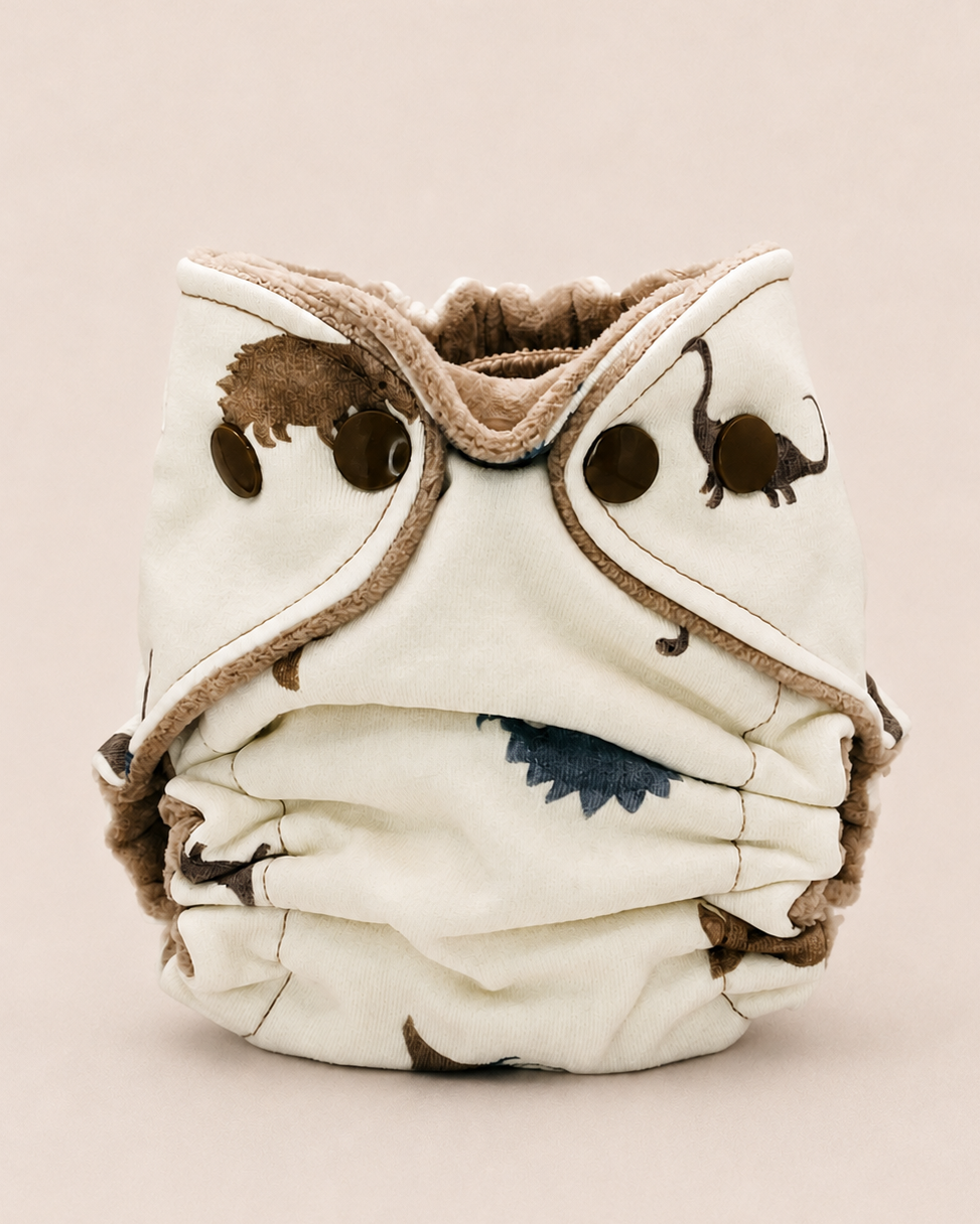 A reusable cloth diaper with a cream-colored fabric body, brown faux-fur trim, and printed brown animal silhouettes, shown against a solid light beige background.