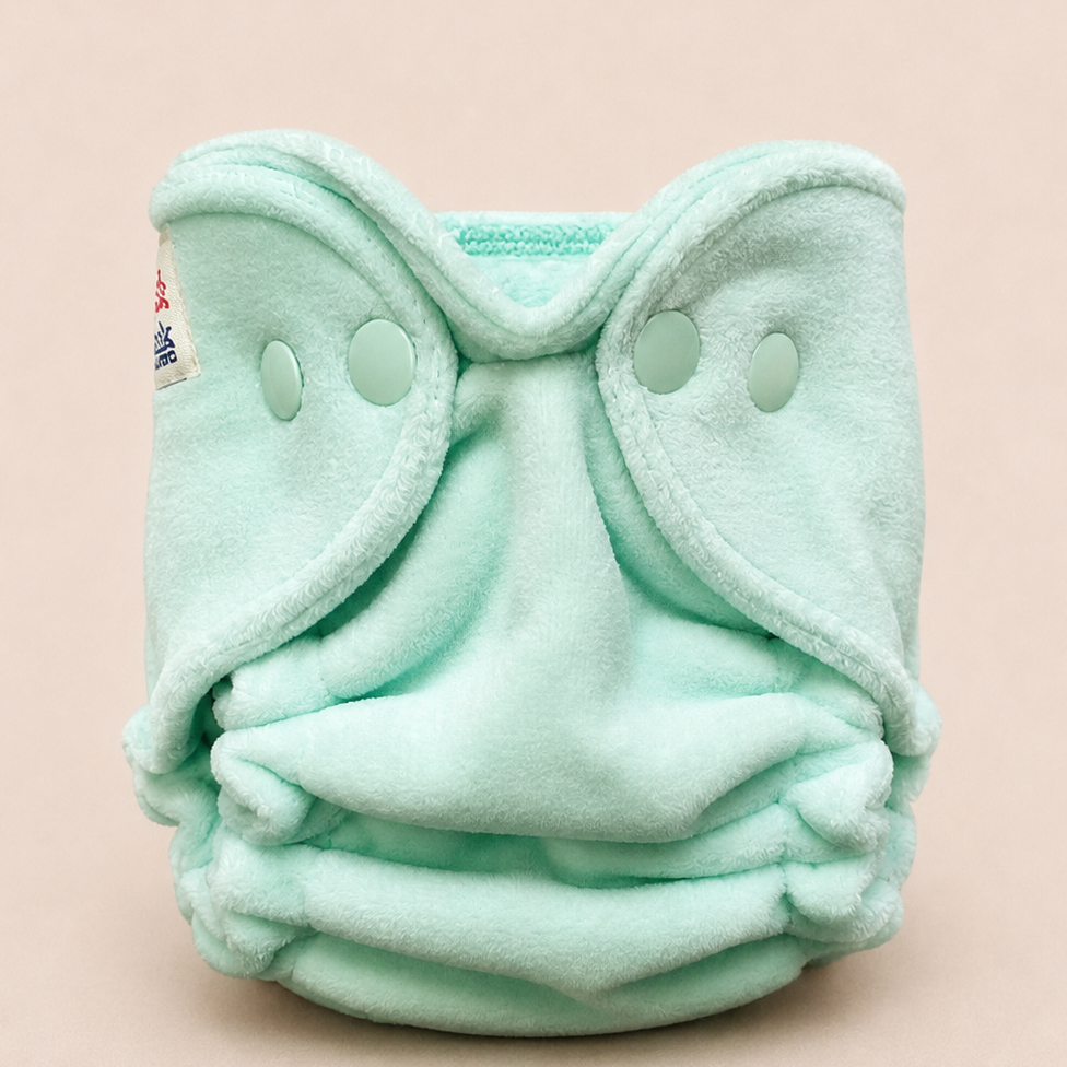 A mint green fleece reusable cloth diaper with snap closures, photographed against a solid pale pink background.