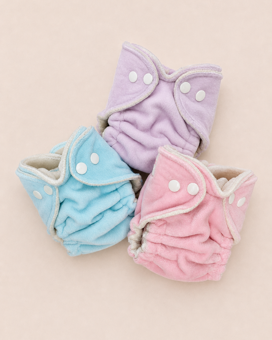 Three reusable cloth diapers in pastel colors, including light blue, lavender purple, and soft pink, arranged on a solid pale peach background.