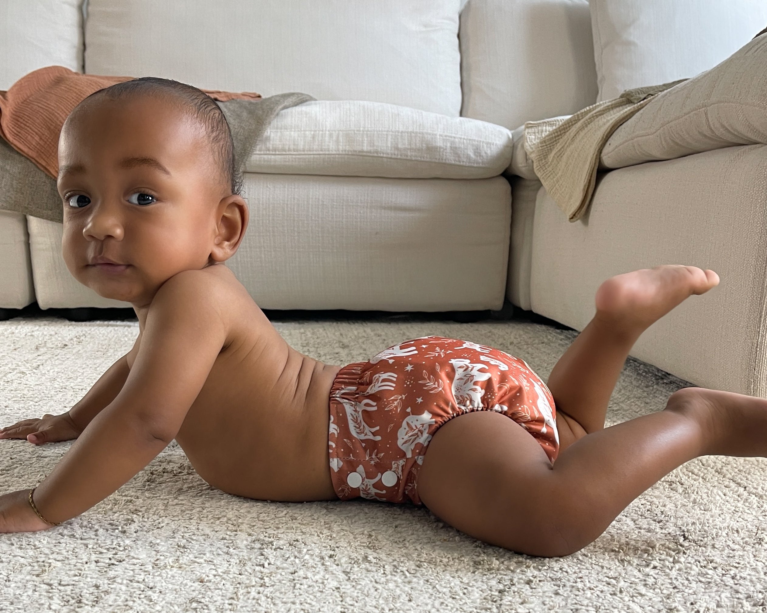 Baby wearing a patterned diaper on a carpeted floor with a beige sofa in the background