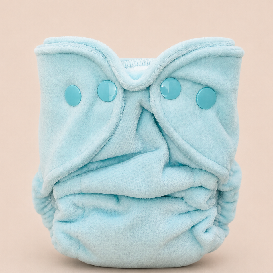 A light blue reusable cloth diaper with three matching snap closures, photographed against a solid pale peach background.