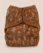 A brown reusable cloth diaper with a beige botanical pattern and brown adjustable snaps, photographed against a solid light beige background.