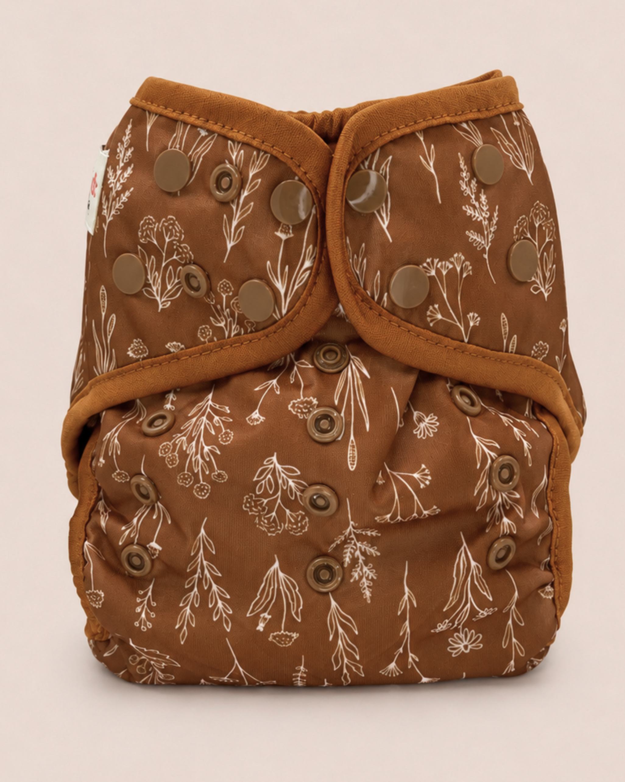 A brown reusable cloth diaper with a beige botanical pattern and brown adjustable snaps, photographed against a solid light beige background.
