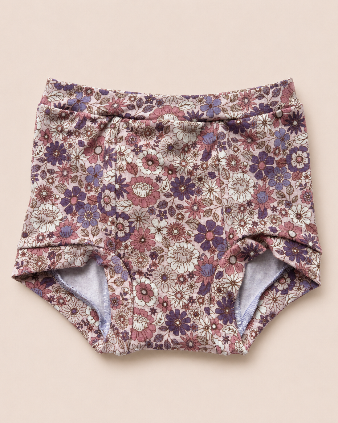 A pair of baby bloomers with a dense floral pattern in dusty pink, purple, and white on a beige background, laid flat on a solid light beige surface.