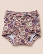 A pair of baby bloomers with a dense floral pattern in dusty pink, purple, and white on a beige background, laid flat on a solid light beige surface.
