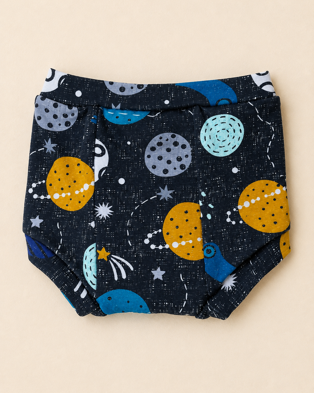 A pair of baby bloomers with a space-themed print featuring planets, stars, and rockets on a dark navy blue textured background, laid flat on a light beige background.