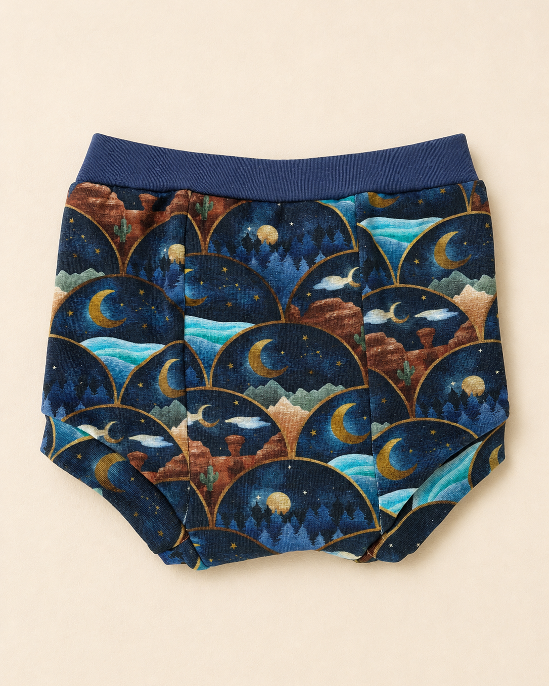 A pair of baby bloomers with a dark blue elastic waistband and a detailed celestial pattern featuring crescent moons, stars, and mountains in shades of navy, blue, brown, and gold, laid flat on a light beige background.