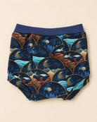 A pair of baby bloomers with a dark blue elastic waistband and a detailed celestial pattern featuring crescent moons, stars, and mountains in shades of navy, blue, brown, and gold, laid flat on a light beige background.