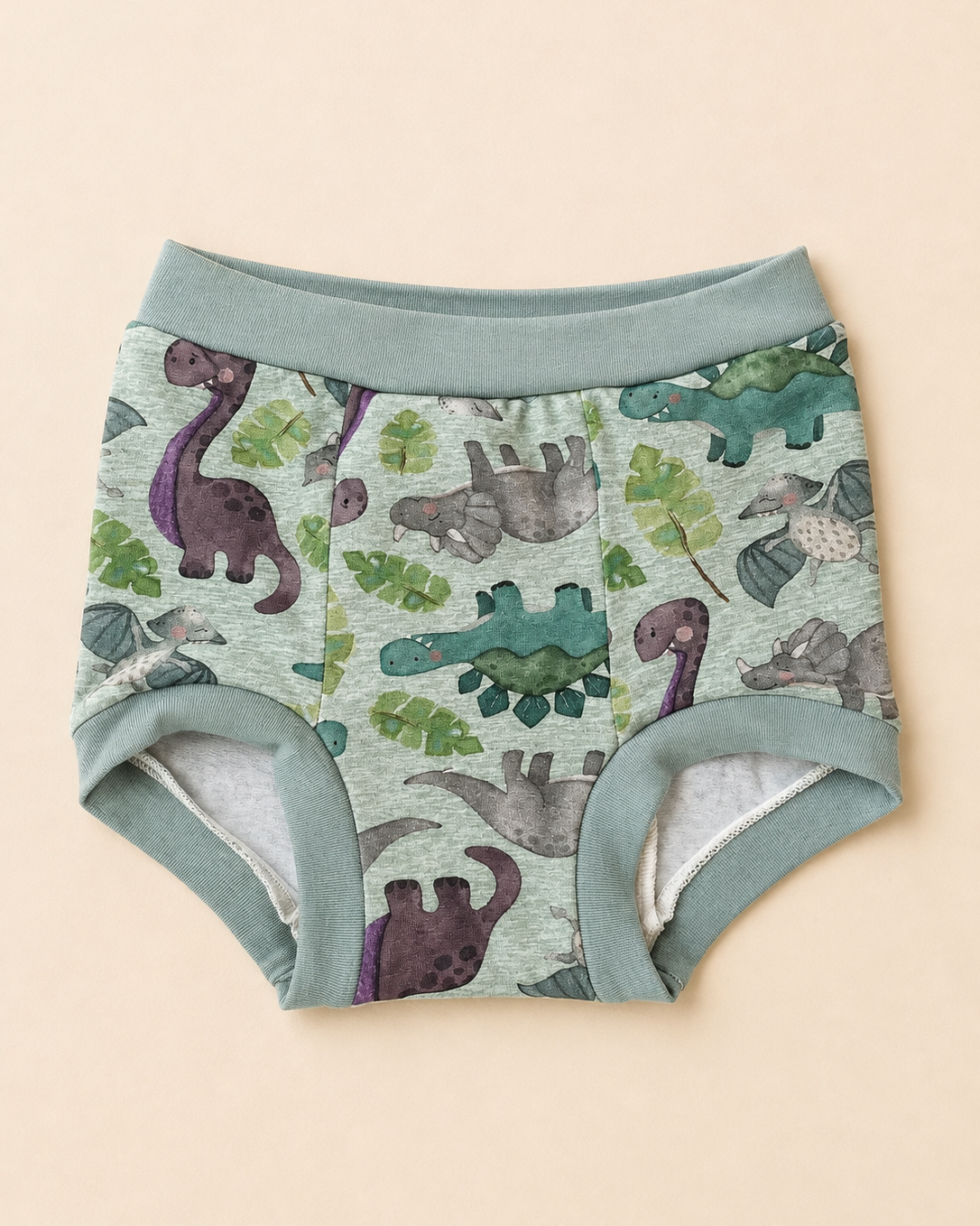 A pair of baby boy's underwear with a green dinosaur and leaf pattern, featuring a solid sage green waistband and leg cuffs, laid flat on a light beige background.