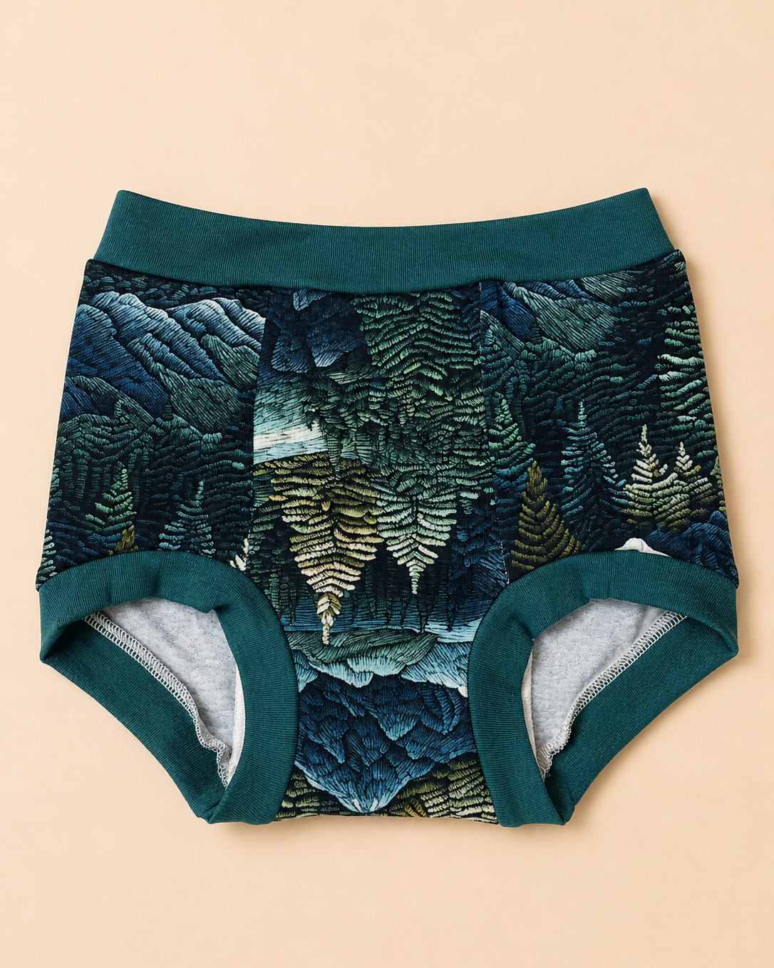 A pair of baby boyshorts featuring a dark teal and forest green fern forest pattern on the exterior, with solid dark teal trim and a light gray heathered interior lining, laid flat on a warm beige background.