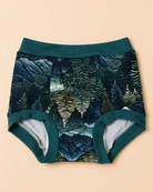 A pair of baby boyshorts featuring a dark teal and forest green fern forest pattern on the exterior, with solid dark teal trim and a light gray heathered interior lining, laid flat on a warm beige background.