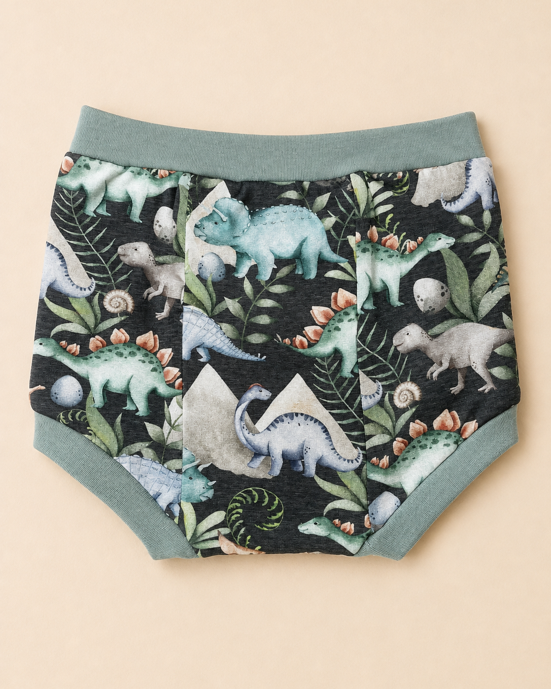A pair of children's diaper cover featuring a dinosaur and prehistoric plant pattern on a black background, with sage green trim and waistband, laid flat on a beige background.