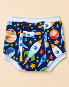 A pair of baby cloth training pants with a colorful space-themed print featuring rockets, planets, and stars on a navy blue background, finished with light blue elastic waist and leg cuffs, shown flat on a beige background.