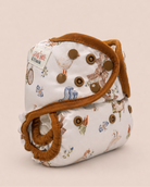 A reusable cloth diaper with a white background and a whimsical multicolor print featuring a chicken, a windmill, and small floral motifs, accented with a brown elastic strap and brown snap closures, set against a solid light beige background.