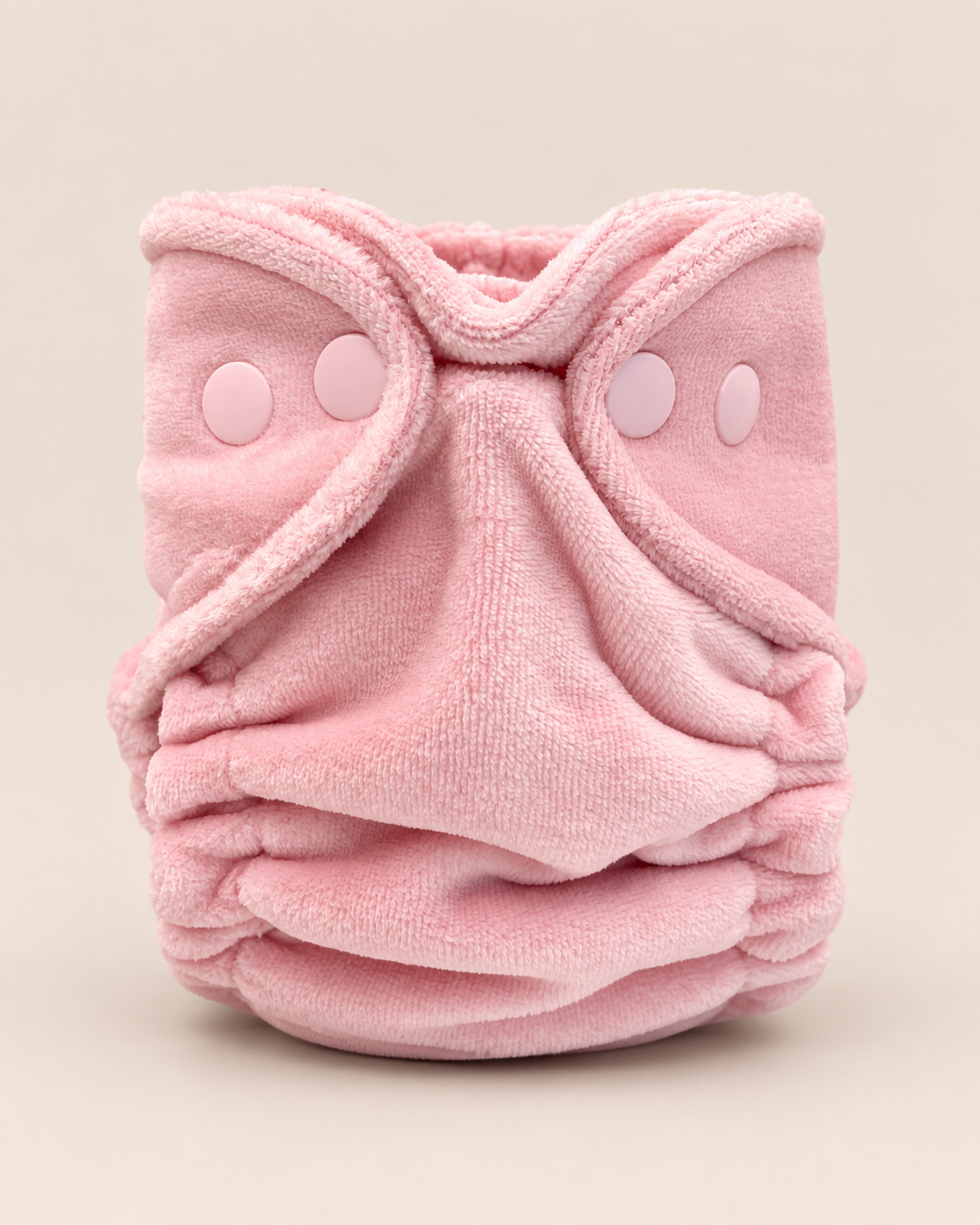 A soft pink reusable cloth diaper with a plush terry cloth texture, featuring adjustable hook-and-loop closure tabs on the front, photographed against a solid light beige background.
