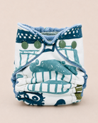 A reusable cloth diaper with a white base and a blue-green nautical print, featuring adjustable straps with green buttons, set against a solid light beige background.