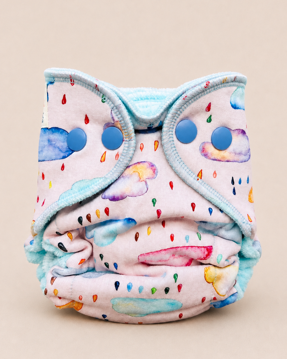 A reusable cloth baby diaper with a light pink background, decorated with a whimsical watercolor cloud and raindrop pattern in multiple colors, featuring a light blue elasticized trim and snap closures.