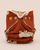 A folded reusable cloth diaper in a rust-orange color with cream trim and three gold-tone snap buttons, photographed on a neutral light beige background.