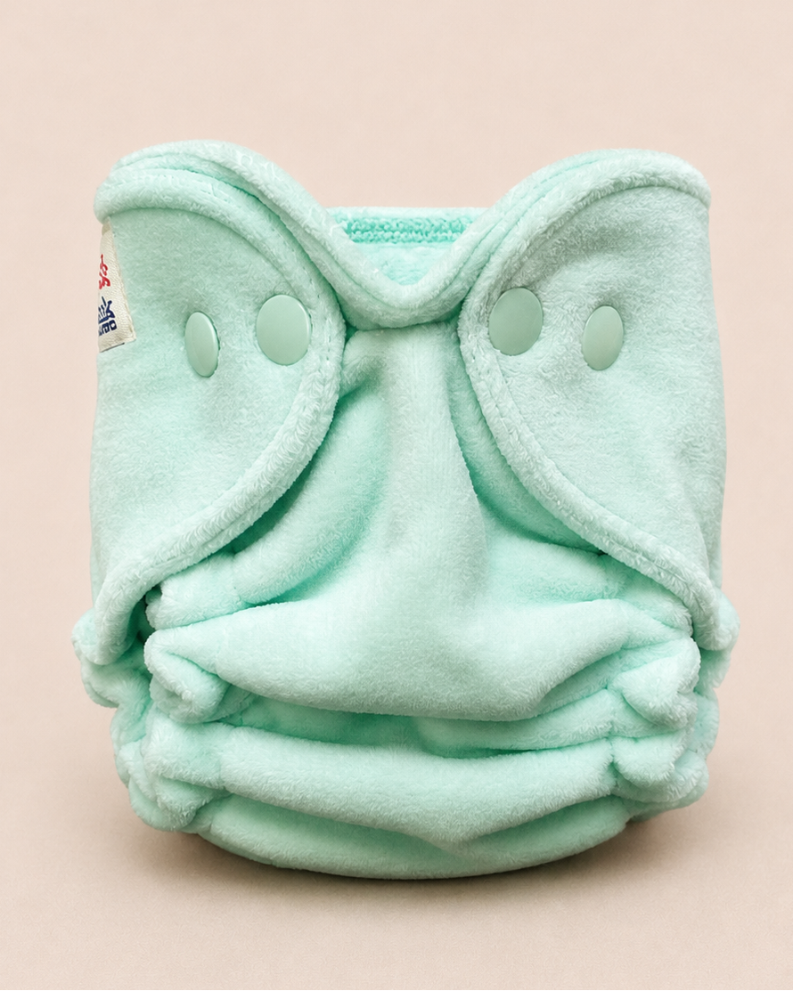 A mint green fleece reusable cloth diaper with snap closures, photographed against a solid pale pink background.