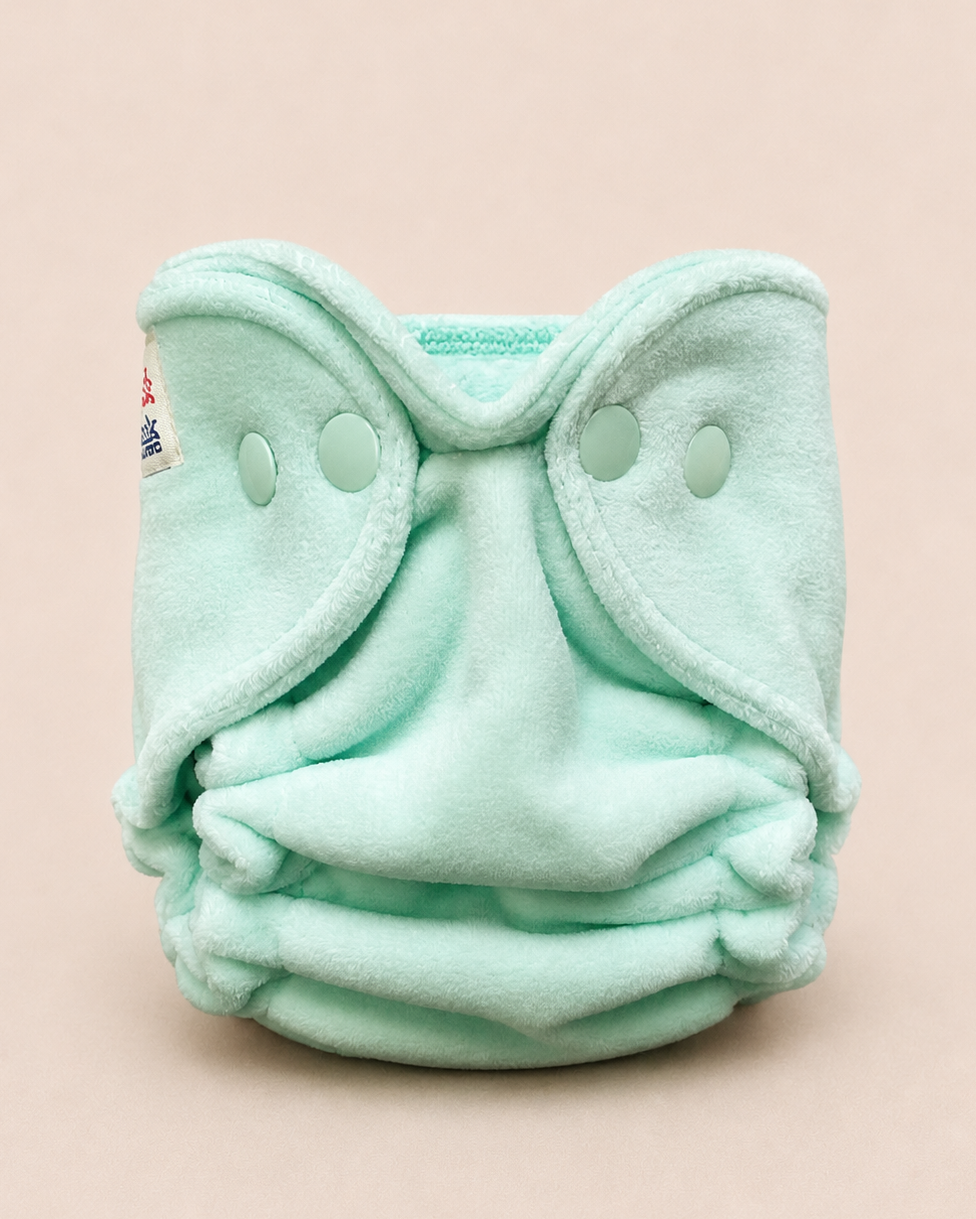 A mint green fleece reusable cloth diaper with snap closures, photographed against a solid pale pink background.
