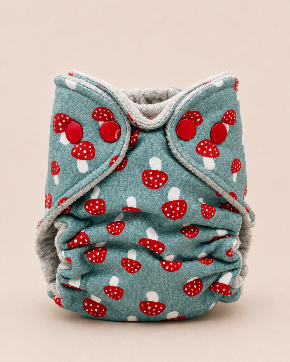 A reusable cloth diaper with a sage green background and a red and white mushroom print, featuring gray fleece trim and a gray inner lining, shown on a solid beige background.