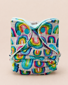 A reusable cloth diaper with a colorful rainbow pattern on a light gray linen-like fabric, featuring a light blue elastic waistband and two turquoise snap closures, photographed against a solid beige background.