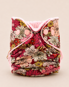 A reusable cloth diaper with a vibrant floral print in shades of red, pink, beige, and green, featuring a soft pink fleece lining and snap closures, set against a solid light beige background.