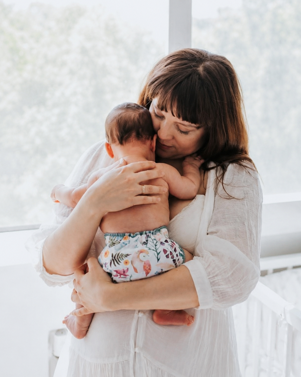A woman in a white long-sleeve dress holds a baby wearing a floral-patterned cloth diaper in a bright, sunlit room.