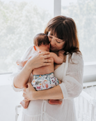 A woman in a white long-sleeve dress holds a baby wearing a floral-patterned cloth diaper in a bright, sunlit room.