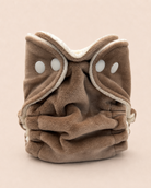 A plush brown cloth face mask with a soft cream fleece trim and two white snap buttons, photographed on a solid light beige background.