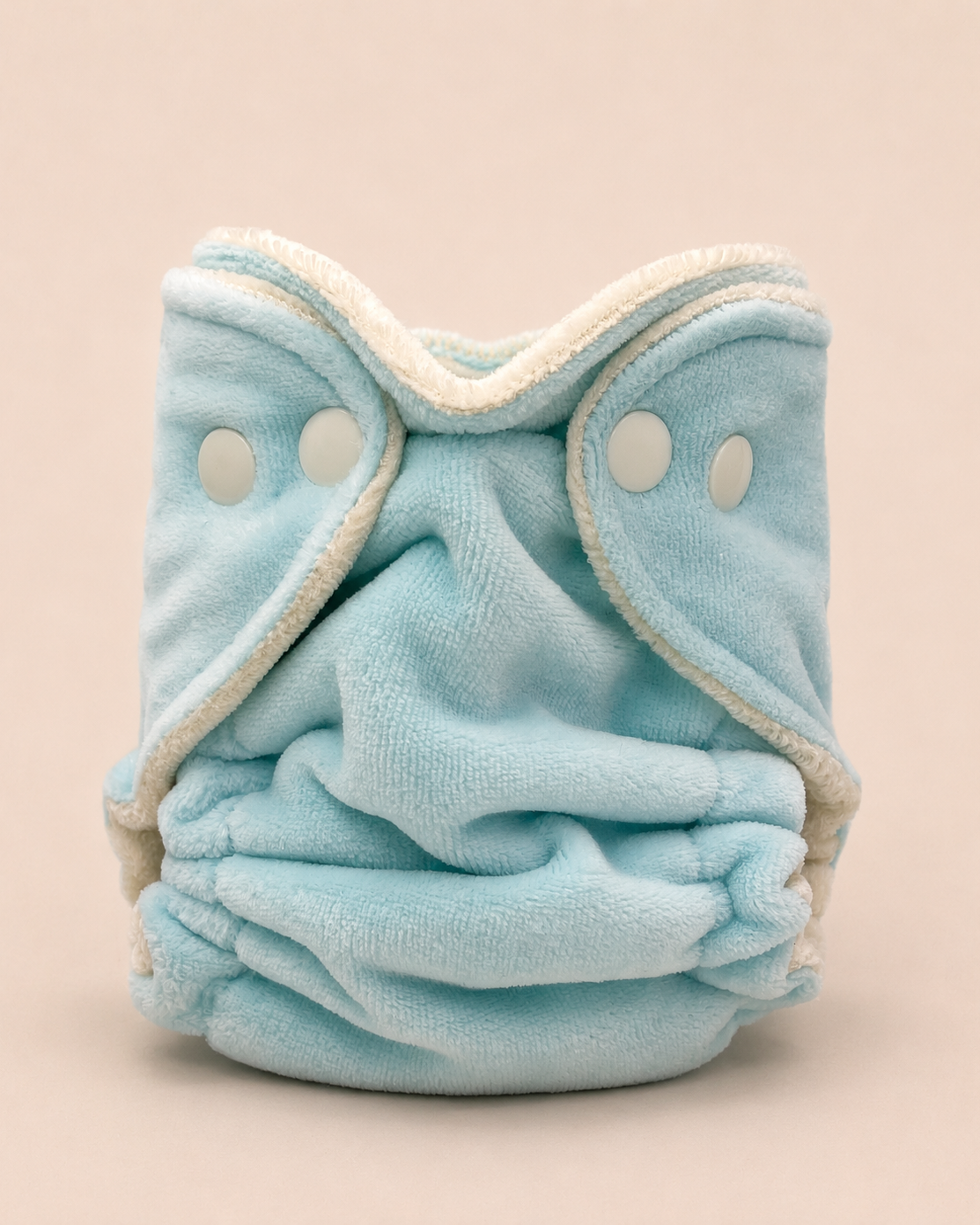 A light blue reusable cloth diaper with cream-colored trim and snap closures, photographed against a solid pale pink background.
