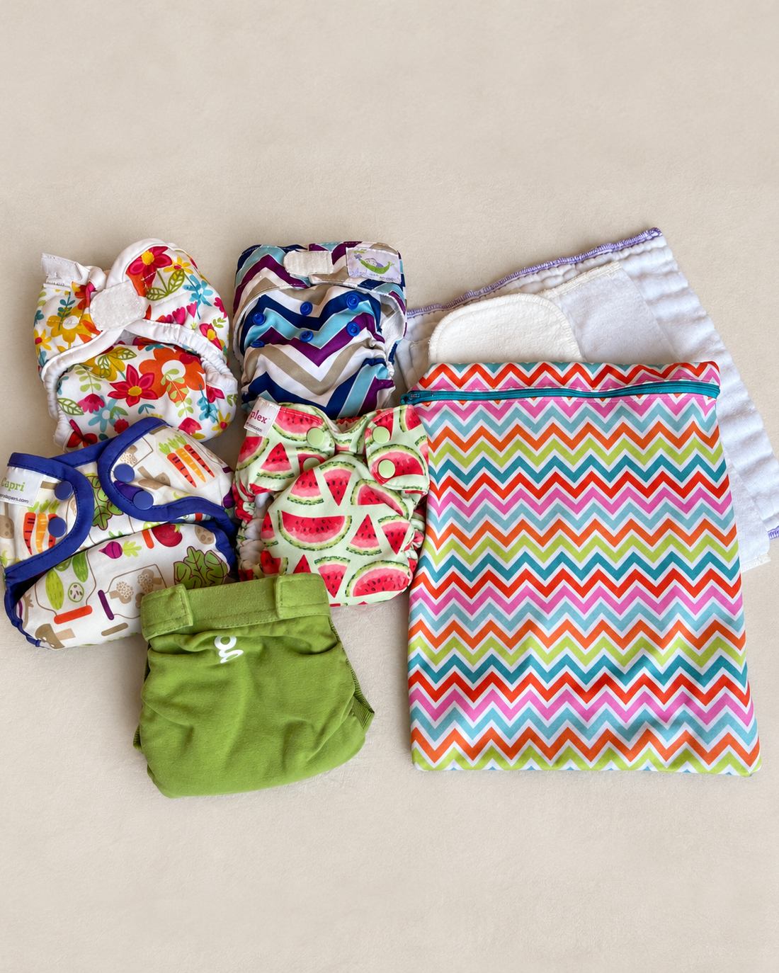 A flat-lay collection of reusable cloth diaper sets and a matching zip pouch on a neutral beige background. The set includes multiple diaper styles with various prints (floral, chevron, watermelon, solid green) and a large zippered pouch featuring a vibrant multicolor chevron pattern.