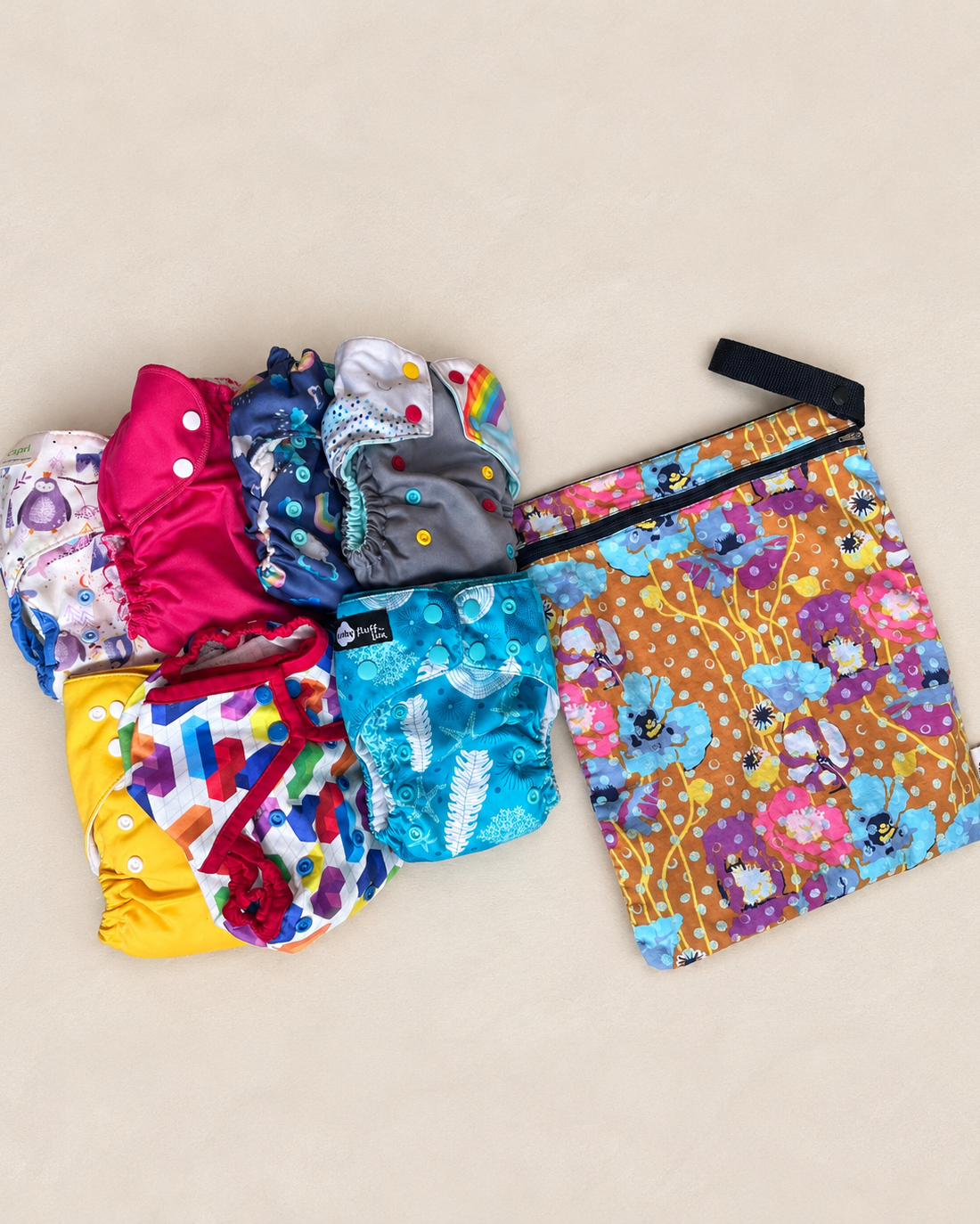 A set of reusable cloth diapers pieces and a matching zip pouch laid out on a light beige background. The set includes several colorful diapers along with a larger zippered pouch featuring a floral and butterfly pattern on a warm orange background.