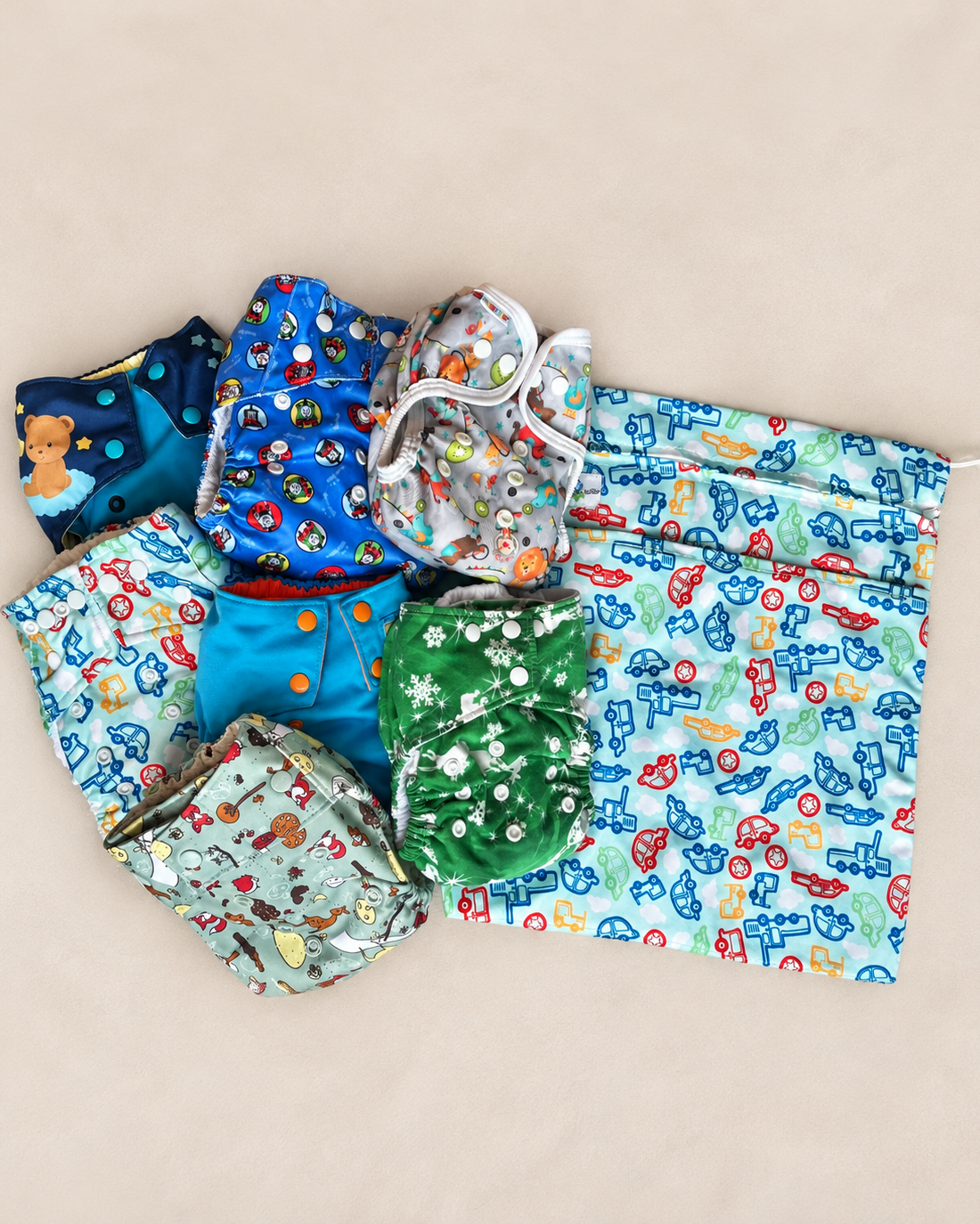 A set of reusable cloth diapers in assorted colorful patterns, including blue, green, and light blue with cartoon and animal prints, arranged on a beige background next to a matching light blue storage bag with a cartoon print.