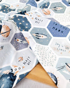 A close-up of patterned fabric with a celestial print, featuring hexagonal panels with stars, planets, rockets, and phrases like 'BLAST OFF' and 'TO THE MOON' on a light wood surface.