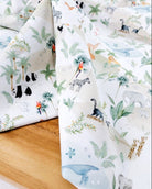 A close-up of a folded white fabric with a colorful safari animal print, draped over a light wooden surface. The print features various animals including zebras, giraffes, elephants, pandas, and a toucan, alongside green palm trees and foliage.