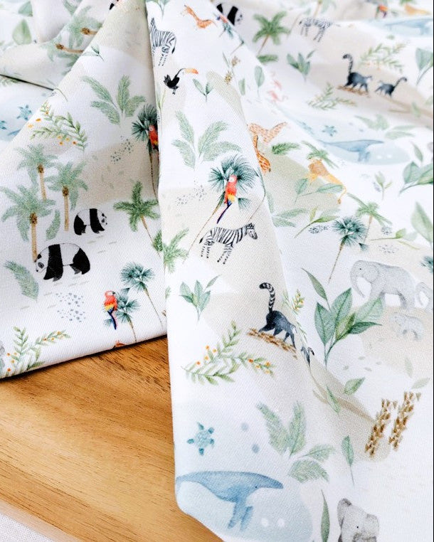 A close-up of a folded white fabric with a colorful safari animal print, draped over a light wooden surface. The print features various animals including zebras, giraffes, elephants, pandas, and a toucan, alongside green palm trees and foliage.
