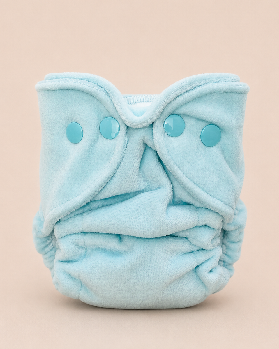 A light blue reusable cloth diaper with three matching snap closures, photographed against a solid pale peach background.