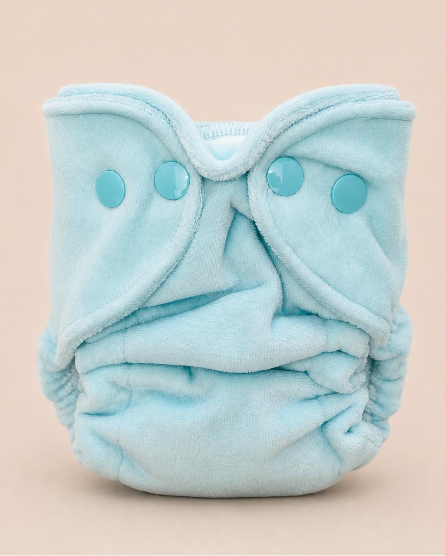 A light blue reusable cloth diaper with three matching snap closures, photographed against a solid pale peach background.